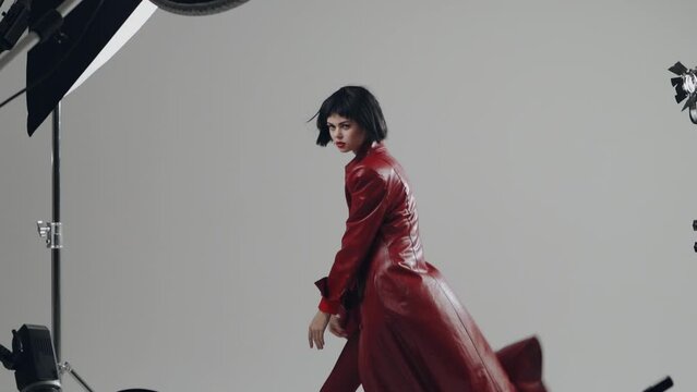 Woman professional fashion model posing in a stylish red coat in the studio for a photo on a white cyclorama with studio fixtures. Posing like a dance