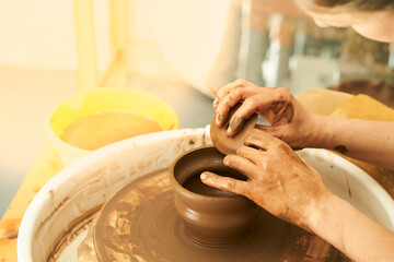 A Potter works with red clay on a Potter's wheel in the workshop..Women's hands create a pot. Girl sculpts in clay pot closeup. Modeling clay close-up. Warm photo atmosphere. 