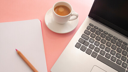 Blank page of a notebook next to a laptop and a white cup with coffee on a light pink background