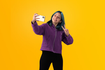 Cheerful nice young girl with blue hair taking selfie on cellphone and smiling. Isolated on yellow baclground - 525699710