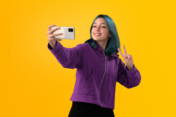 Cheerful nice young girl with blue hair taking selfie on cellphone and smiling. Isolated on yellow baclground - 525699715