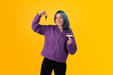 Smiling young woman student with blue hair holds a credit card and key over a yellow background - 525699720