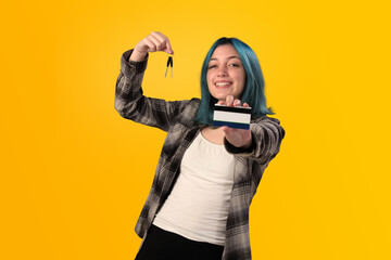 Smiling young woman student with blue hair holds a credit card and key over a yellow background - 525699729