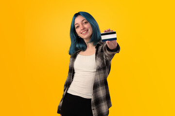 Smiling young woman student with blue hair holding a credit card over yellow background - 525699732