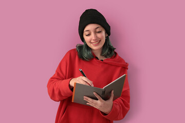 Cute young girl with blue hair taking notes over a pink background - 525699987