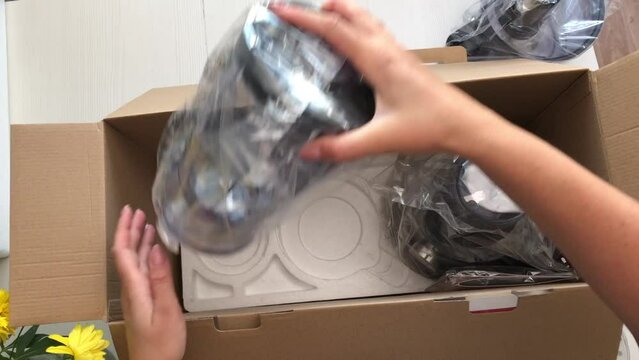 A woman opens a box with a food processor. He takes out the packed components of the combine from it and puts it on the table. Shot from above, close-up