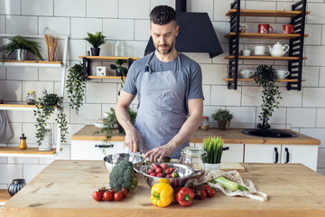 Handsome father, strong young man cooking healthy vegetable salad with fresh organic ingredients, tasty food in the kitchen at home . Men doing chores. Ripe pepper, tomato, cucumber