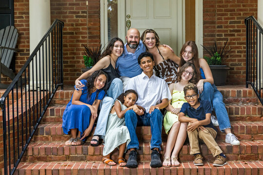 A large and blended and mixed happy family sitting on the front steps of their brick home in the suburbs