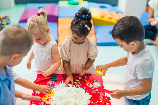 Fun sensory play with moldable and fluffy kinetic sand at nursery school. Toddlers standing around the table and using different tools for sculpting sand such as colorful and textured rolling pins
