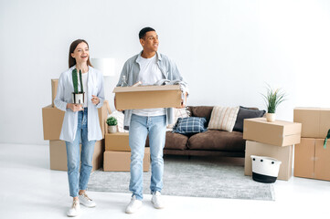 Fototapeta na wymiar Moving to a new apartment. Happy multiracial young couple in love walk into their new home, holding boxes of household items, look around the interior with delight, rejoice at their new home, smile