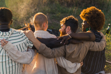 Rear view of multicultural good old friends hugging and having a weekend picnic in nature near the campfire, talking and laughing. Sunset time. High quality photo
