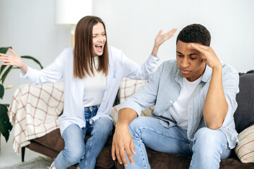 Misunderstanding in relationships, jealousy, quarrel. A married multiracial couple sorts things out while sitting in the living room on a sofa, a caucasian woman screams at her hispanic husband