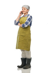 gardening, farming and old people concept - smiling senior woman in green garden apron and rubber boots calling on smartphone over white background