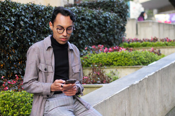 young man with glasses using his cell phone while sitting in a park sending a message