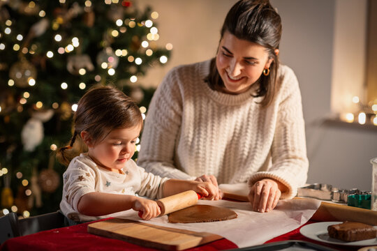 family, cooking and winter holidays concept - happy mother and baby daughter with rolling pin making gingerbread cookies from dough at home on christmas