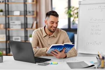distant education, school and remote job concept - happy smiling male math teacher with laptop computer, book and flip chart having online class or video call at home office