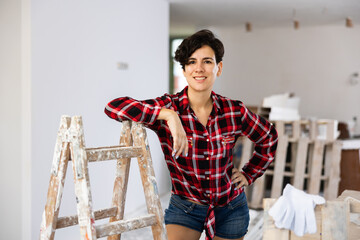Attractive female builder standing and leaning at stepladder in apartment during repair works