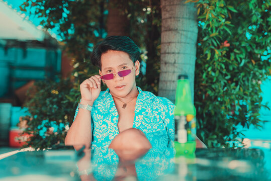 Asian Man In An Unbuttoned Hawaiian Luau Polo Shirt And Necklace Holding His Purple Sunglasses Near A Reflective Glass Table In A Garden.