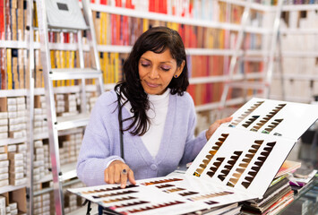 Positive hispanic woman visiting specialized store of hair cosmetics, choosing dye for coloring.