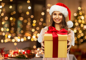 Fototapeta na wymiar christmas, holidays and people concept - happy smiling teenage girl in santa helper hat holding gift box over festive lights at home on background