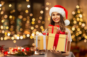 christmas, holidays and people concept - happy smiling teenage girl in santa helper hat holding gift box over festive lights at home on background