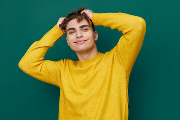 a relaxed man with curly hair, standing on a green background in a yellow sweater and closing his eyes holding his head with his hands