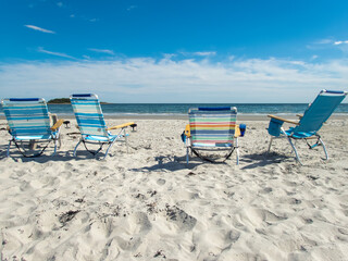 Four empty beach lounge chairs in the sand with shoreline and sky in the background.
