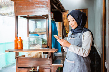 woman wearing a veil wearing an apron using a cellphone while selling at a chicken noodle cart