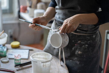 Experienced woman in black apron enjoys working with handmade craft in pottery studio. Female artisan uses brush to paint white ceramic cup with creative pattern standing on blurred background closeup