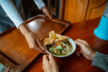 waiter's hand serving a bowl of chicken noodle dish to customers on the wood table