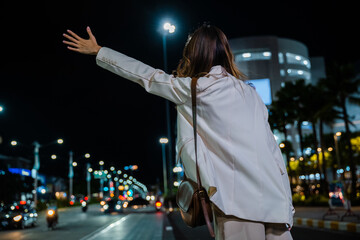 Back view, Beautiful woman smiling using smartphone application hailing with hand up calling cab outdoor after late work, Asian business woman hail waving hand taxi on road in city street at night