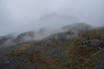 Dark atmospheric landscape in fading autumn colors with high rocky hill and silhouette of sharp rocks on mountain top in dense fog in rainy weather. Large black peak in thick low clouds during rain.