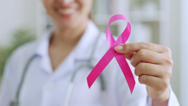 Smiling young Asian doctor woman hold pink ribbon as sign of October Breast Cancer Awareness month