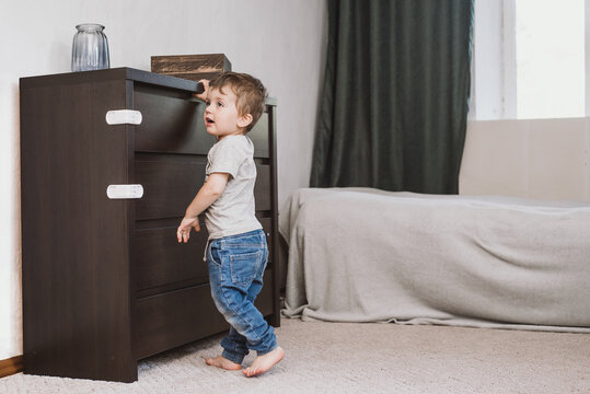 Little Boy Two Years Old Trying To Open Chest Of Drawers With Safety Latches. Child Unsupervised Plays With Cabinets. Danger For The Baby To Pinch The Hand Of The Cabinet Door.