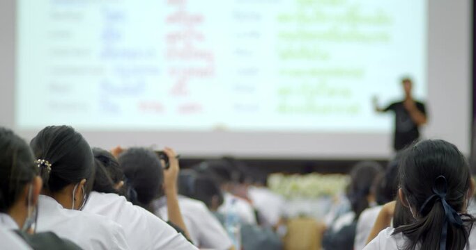 Slow motion scene, students in white uniforms are sitting in lecture halls in a large auditorium and raise their hands to answer the questions. A teacher stands in front of a large projector screen.