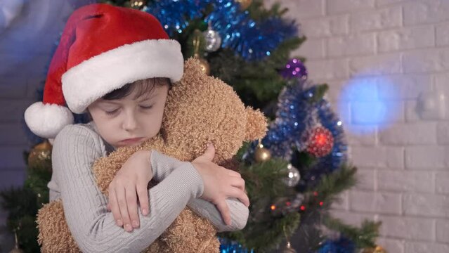 Desperate child with teddy at Christmas. A sad little girl embrace her teddy bear by the Christmas tree. A concept of lonely New Year holidays.