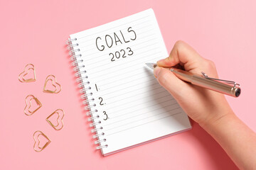Female hand writes goals 2023 in a notebook on a pink background close-up.