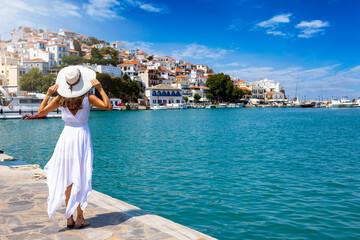 A beautiful tourist woman in a white summer dress enjoys the view to the city of Skopelos island,...