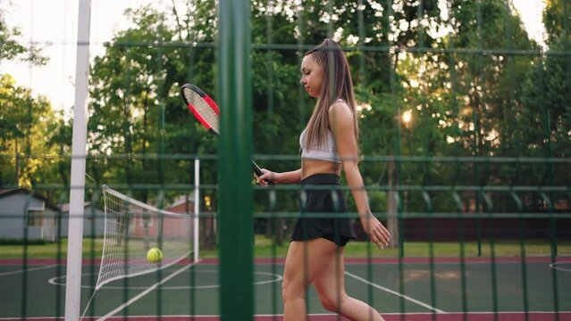 pretty young female tennis player is walking on empty court and bouncing ball by racket
