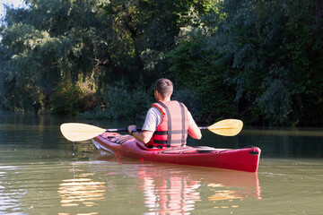 Man kayaking on red kayak in the summer river near green trees