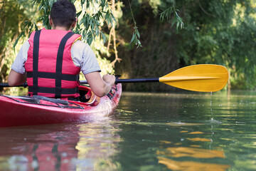 Close up of rear view of man kayaking on red kayak in the summer river near green trees