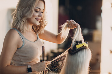 Hairdresser female making hair extensions to young woman with blonde hair in beauty salon....