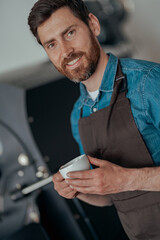 Portrait of male barista with cup of fresh coffee on background of roasting machine