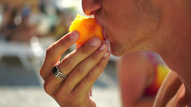 Man eating peach. Young man in sunglases eating peach on the beach. Happy man is smiling and resting at sea. Guy enjoying fruits on summer vacation. Vegetarian and healthy lifestyle concept.