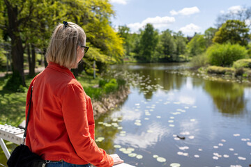 Smiling beauty Caucasian Young Adult Woman with Sunglasses in King's Botanical Garden in Copenhagen, Denmark Wearing Red Jacket and Jeans with Park Pond looking at camera. 