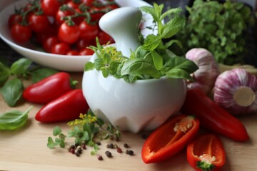 Mortar with fresh herbs near garlic, pepper and cherry tomatoes on wooden table, closeup