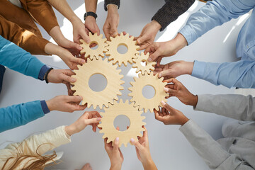 Business people or office workers hold wooden gears that symbolize well-coordinated teamwork. Top view close up of hands of multiracial men and women standing in circle. Concept business cooperation - obrazy, fototapety, plakaty