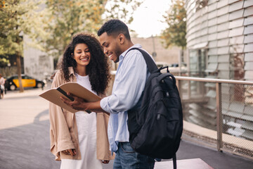 Positive young african classmates with notebook getting ready for class standing outdoors. Brunettes guy and girl wear casual clothes and backpack. Mood, lifestyle, concept.