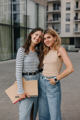 Stylish young caucasian girls hugging posing looking at camera outdoors. Brunette and blonde wear casual clothes in spring. Relaxed lifestyle, concept
