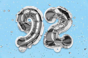 Silver foil balloon number, digit ninety two on a blue background with sequins. Birthday greeting card with inscription 92. Top view. Numerical digit. Celebration event, template.
